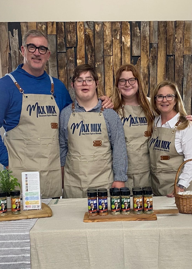 A picture of Max at a product demo with his parents and sister. All are wearing Max Mix branded aprons and smiling into the camera.