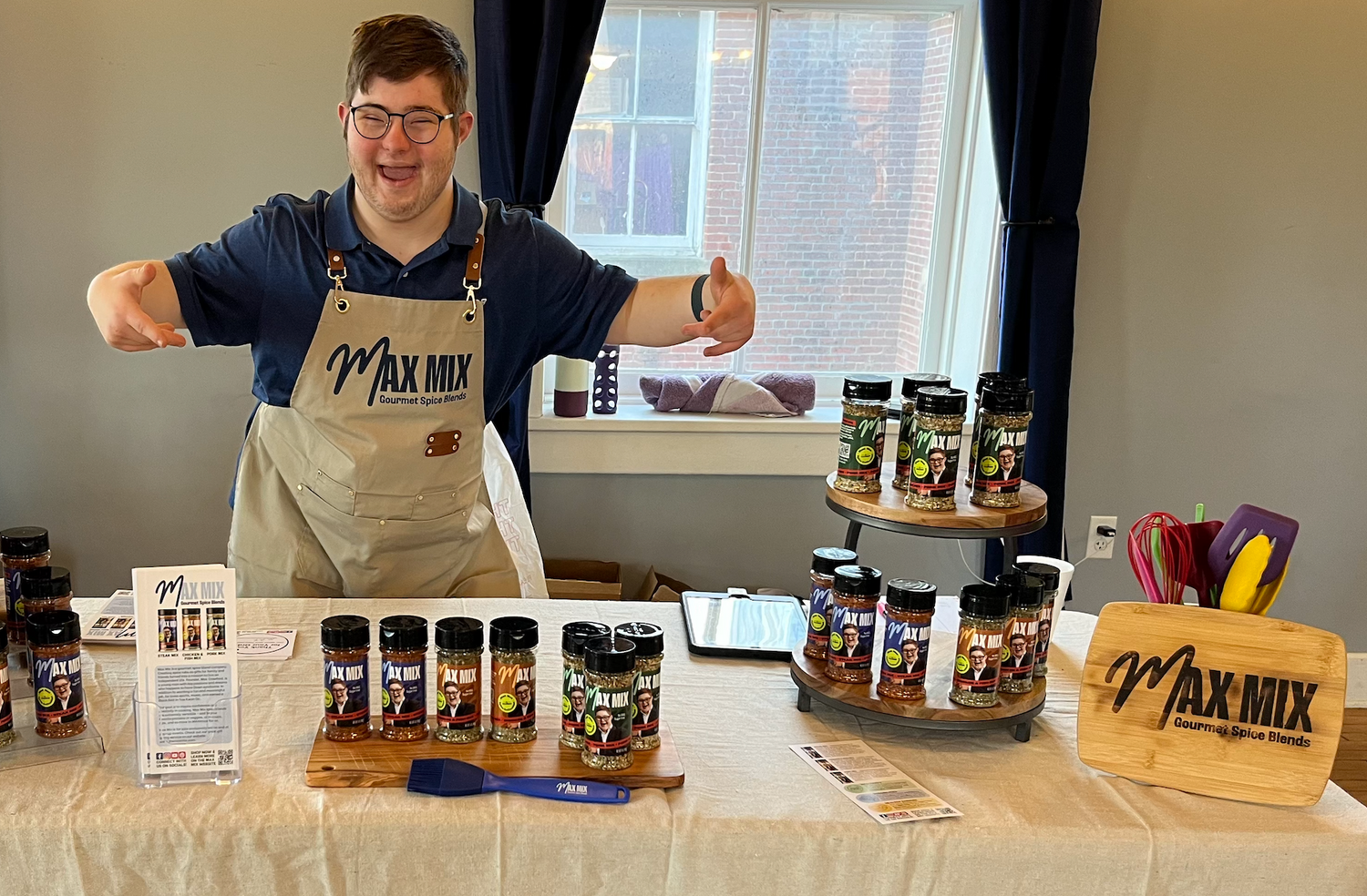 A picture of Max Mix founder Max at a demo, wearing a Max Mix branded apron, enthusiastically pointing at the Max Mix shakers on the table and smiling.