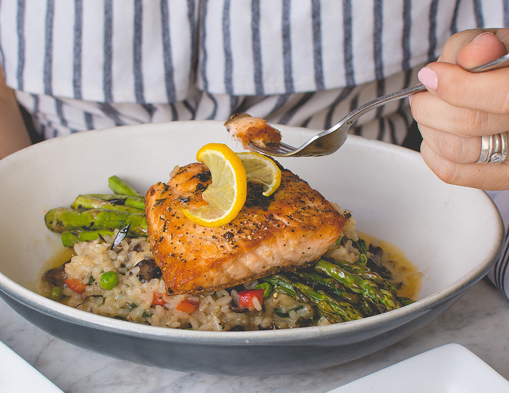 A serving dish full of salmon, rice, and vegetables. A hand holding a fork scoops up a forkful of salmon.