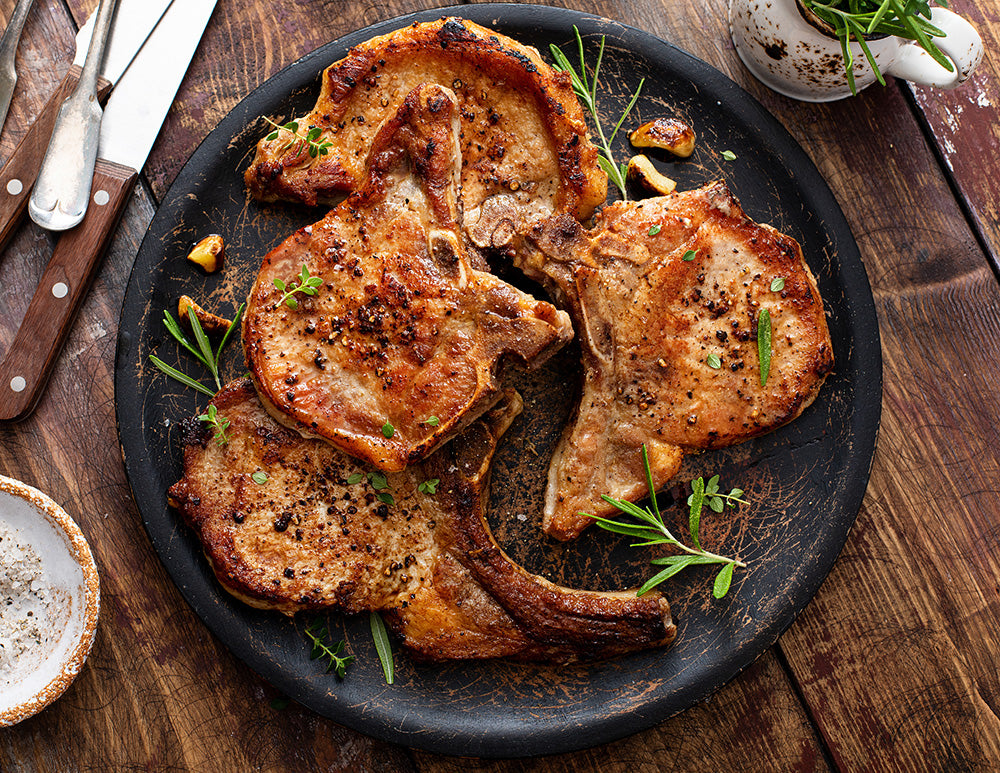 A serving dish full of pork chops, with garlic and fresh herbs, sitting on a wooden table.