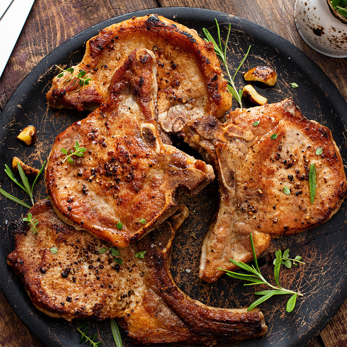 A serving dish full of pork chops, with garlic and fresh herbs, sitting on a wooden table.