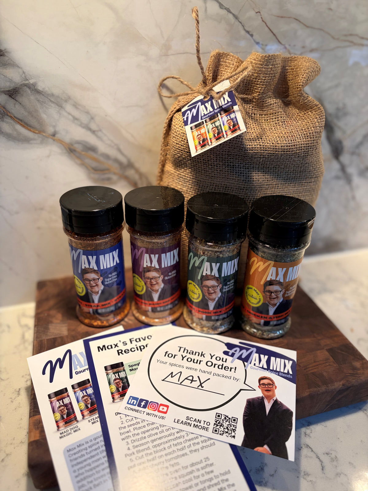 Four bottles of Max Mix product (Steak Mix, Pork Mix, Chicken & Fish Mix, and Mad Dog BBQ), arranged on a wooden cutting board with a burlap drawstring bag and printed cards, set against a marble-patterned background.