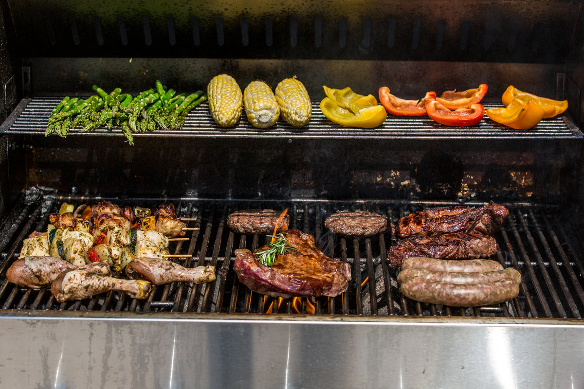 A full grill of cooking steak, burgers, sausage, skewers, chicken wings, and veggies.