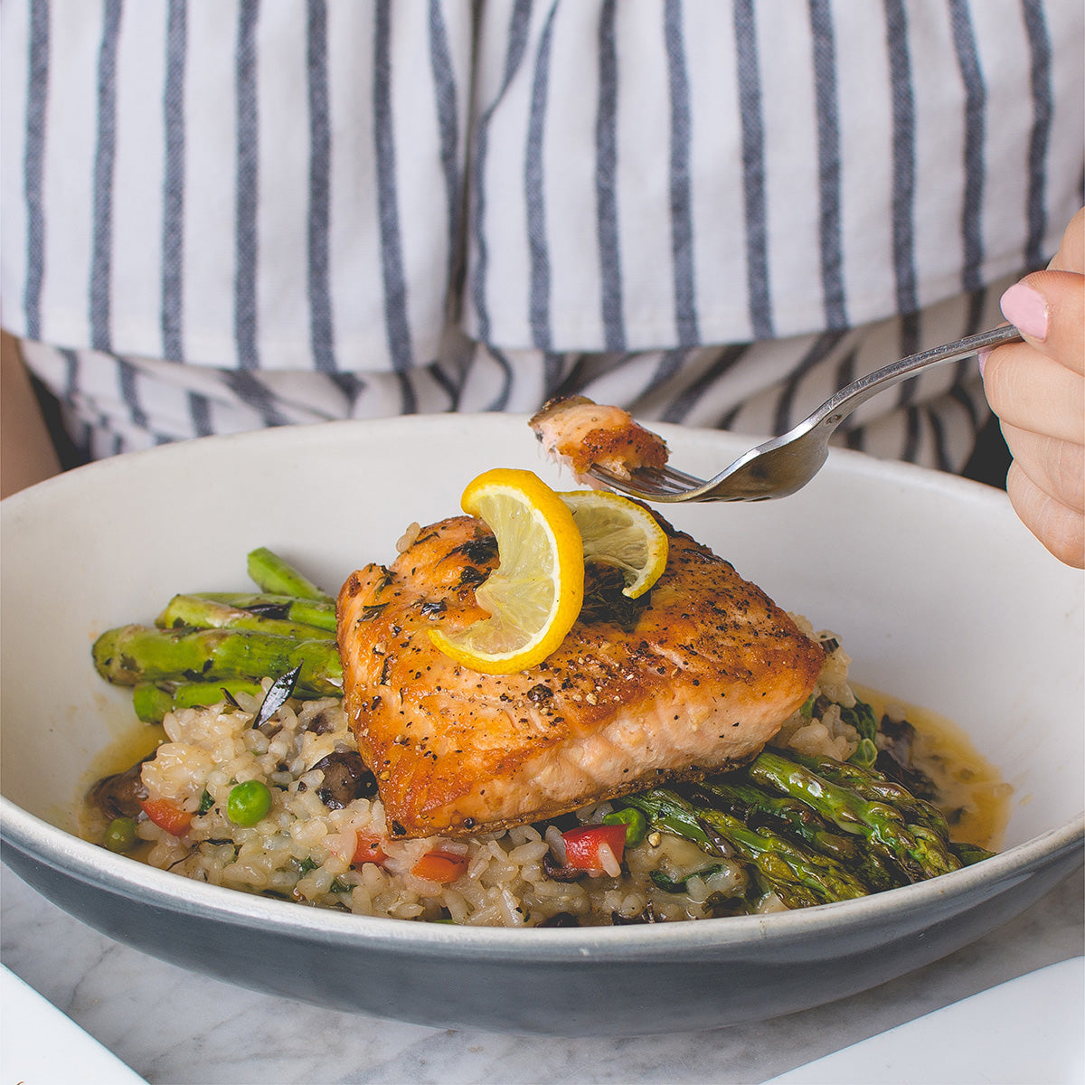 A serving dish full of salmon, rice, and vegetables. A hand holding a fork scoops up a forkful of salmon.